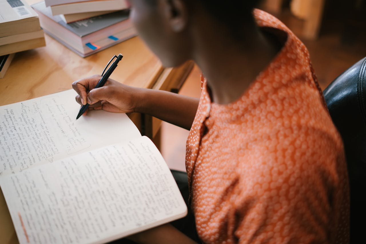 services-03 Focused black woman writing in a notebook with stacked books on a desk, illustrating learning.