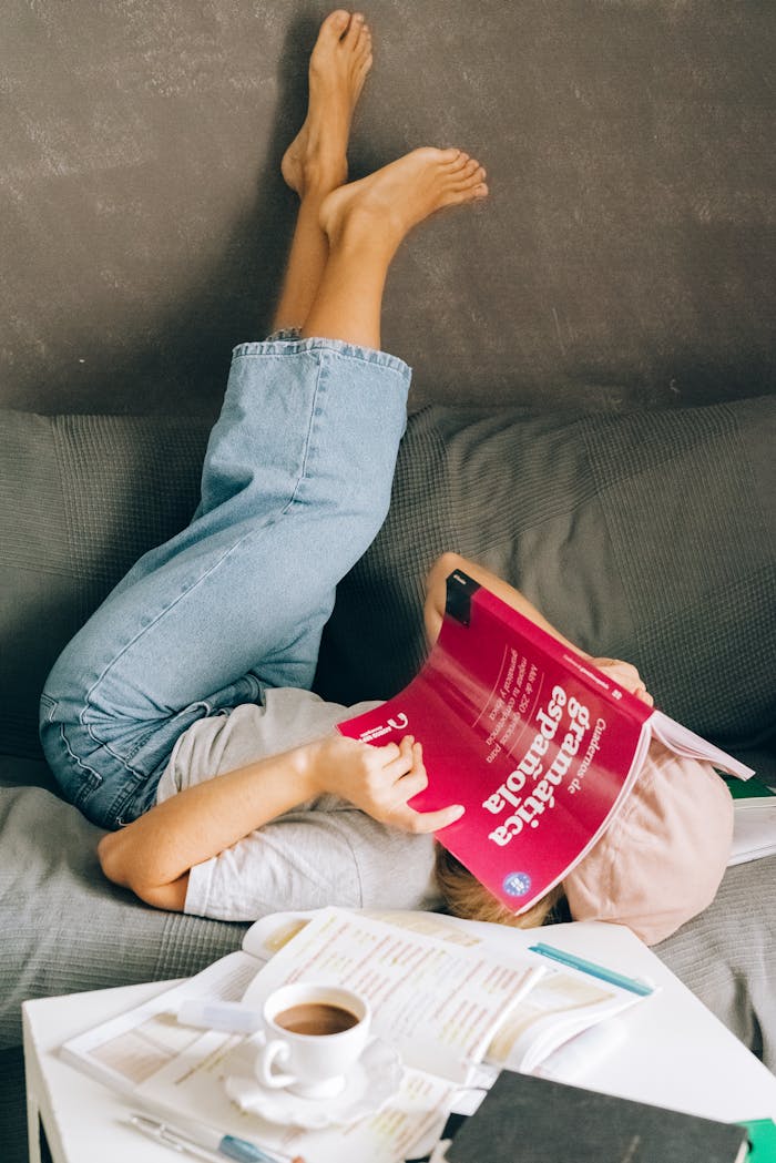 services-02 Young woman relaxes upside-down on a couch while studying a Spanish book indoors.